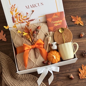 May include: A gift box containing a beige knit blanket tied with an orange ribbon, a Goodio Pumpkin Spice chocolate bar, a round woven coaster, a gold spoon, a cream mug, a small jar, a wooden honey dipper, and a decorative orange pumpkin.