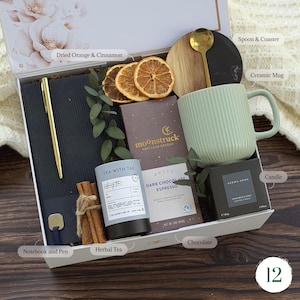 May include: A gift box containing a black notebook with a gold pen, a light green ribbed ceramic mug, a gold spoon, a round wood and black coaster, a Moonstruck dark chocolate espresso bar, a Tea With Tae herbal tea tin, and a candle.