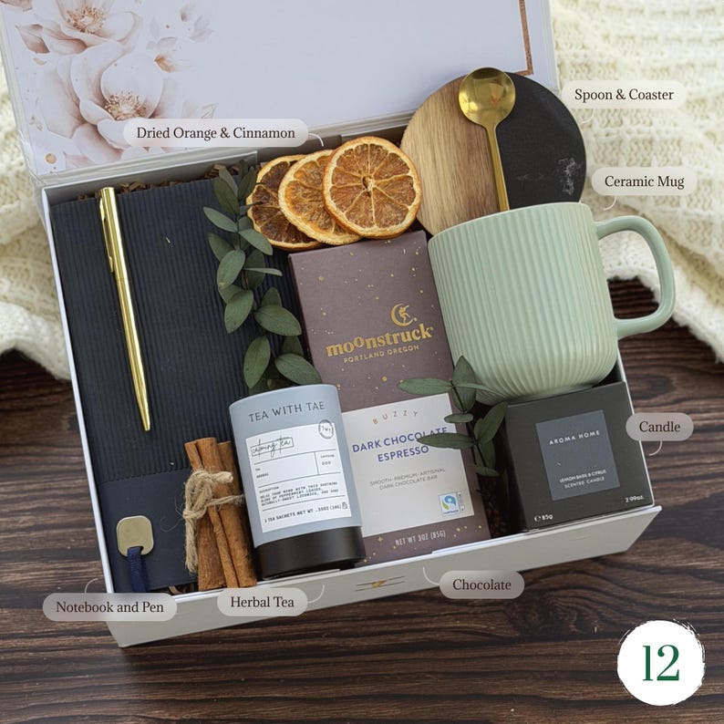 May include: A gift box containing a black notebook with a gold pen, a light green ribbed ceramic mug, a gold spoon, a round wood and black coaster, a Moonstruck dark chocolate espresso bar, a Tea With Tae herbal tea tin, and a candle.