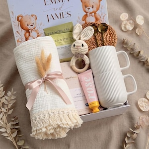 May include: A gift box with baby-themed items. Includes a white blanket with a pink ribbon, two white mugs, a crochet bunny rattle, a chocolate bar, and tea packets. The box is decorated with teddy bears and the names "Emma and James".