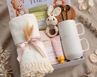 May include: A gift box with baby-themed items. Includes a white blanket with a pink ribbon, two white mugs, a crochet bunny rattle, a chocolate bar, and tea packets. The box is decorated with teddy bears and the names "Emma and James".