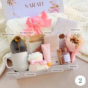 May include: A gift box filled with self-care items. Includes a white ceramic mug, cozy socks, shea butter gloves, hand cream, chocolate, bath salts, and a dried flower bouquet. A pink bow adorns the box, with the name "SARAH" visible.