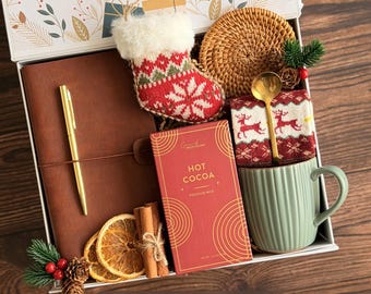 May include: A gift box containing a brown leather journal with a gold pen, a red and white knit stocking ornament, a round woven coaster, a red box of hot cocoa, a gold spoon, a green ribbed mug, dried orange slices, and cinnamon sticks.
