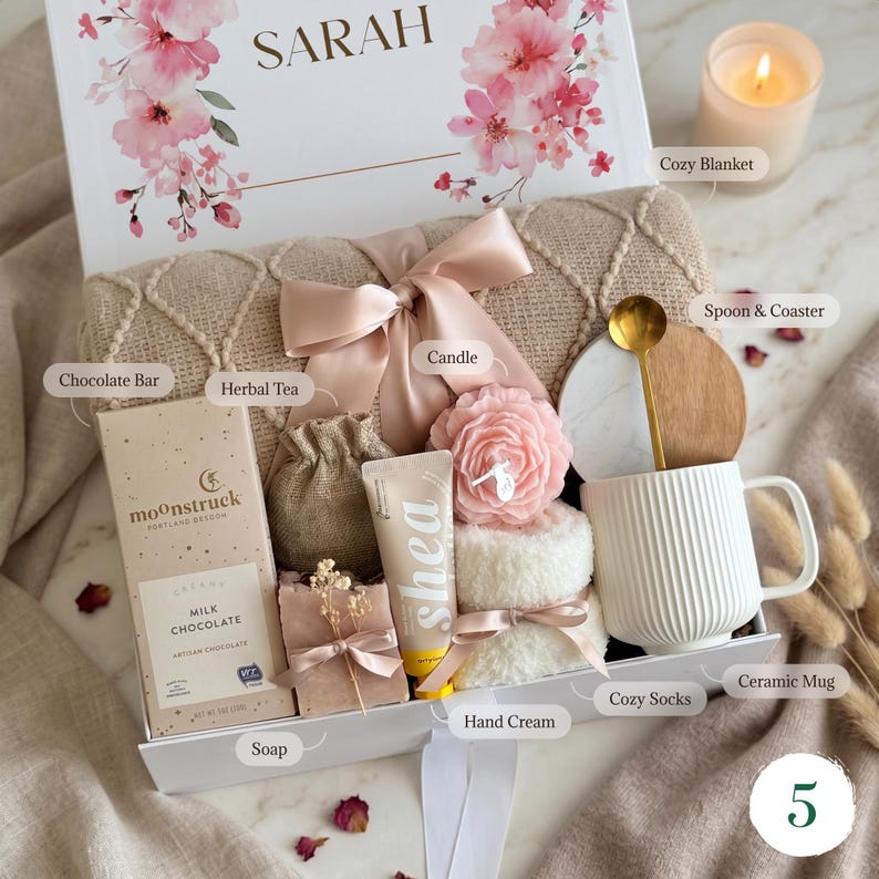 May include: A gift box with a floral design and the name "SARAH" on the lid. Inside are a cozy blanket, candle, chocolate bar, herbal tea, soap, hand cream, cozy socks, a ceramic mug, and a spoon with a coaster. The box is tied with a ribbon.