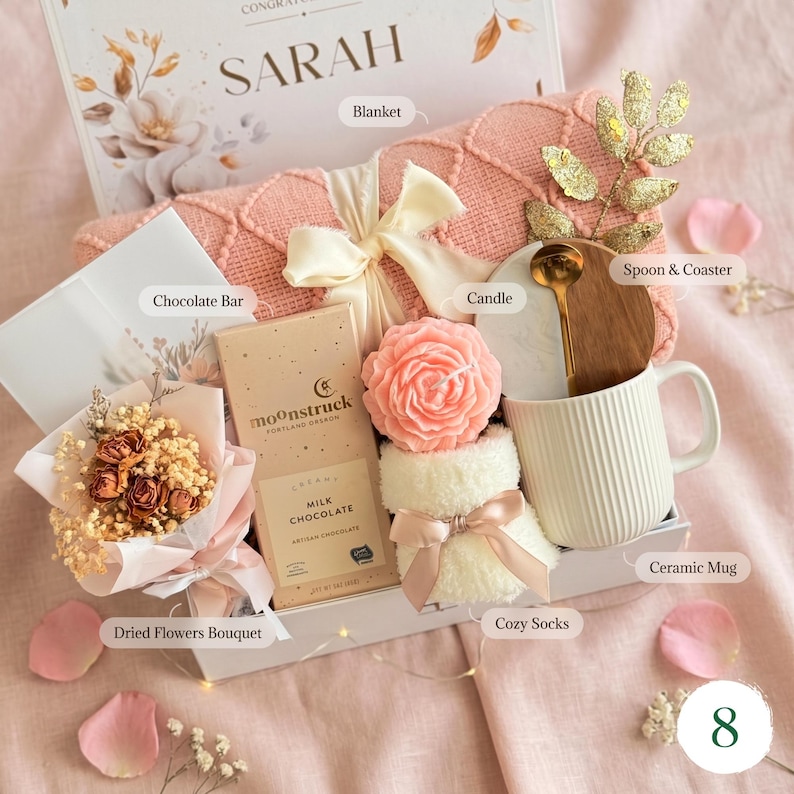 May include: A gift box with a pink blanket, a white ceramic mug, a chocolate bar, a dried flower bouquet, a rose-shaped candle, cozy socks, and a wooden spoon. The box is decorated with a bow and the name "SARAH".