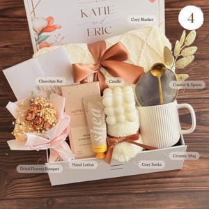 May include: A gift box filled with a cozy blanket, candle, ceramic mug, chocolate bar, hand lotion, dried flowers, and cozy socks. The box is tied with a brown ribbon and includes a coaster with a spoon. The box is labeled "KATIE and ERIC".