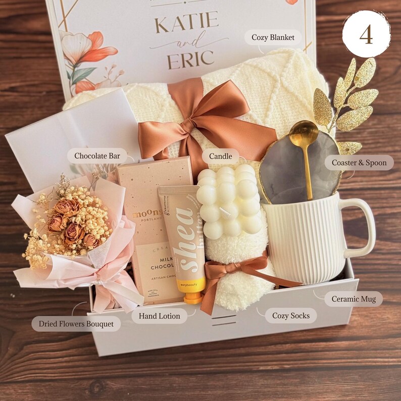 May include: A gift box filled with a cozy blanket, candle, ceramic mug, chocolate bar, hand lotion, dried flowers, and cozy socks. The box is tied with a brown ribbon and includes a coaster with a spoon. The box is labeled "KATIE and ERIC".
