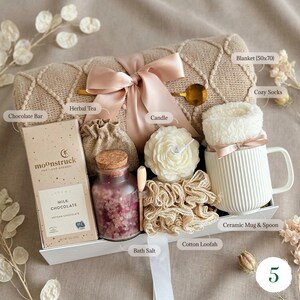 May include: A gift set containing a beige textured blanket with a satin ribbon, a Moonstruck milk chocolate bar, a small burlap pouch, a glass jar of pink bath salts with a cork lid, a flower-shaped candle, a cotton loofah, and a white ribbed ceramic mug.