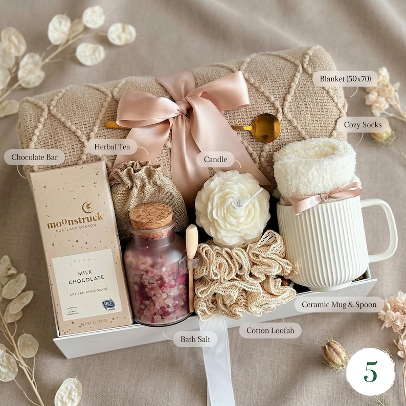 May include: A gift set containing a beige textured blanket with a satin ribbon, a Moonstruck milk chocolate bar, a small burlap pouch, a glass jar of pink bath salts with a cork lid, a flower-shaped candle, a cotton loofah, and a white ribbed ceramic mug.
