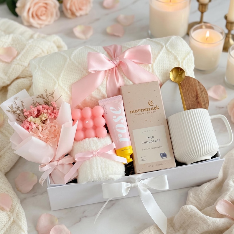 May include: A gift box filled with a white blanket, pink bow, pink candle, chocolate bar, mug, and a bouquet of dried flowers. The box is decorated with pink rose petals and candles.