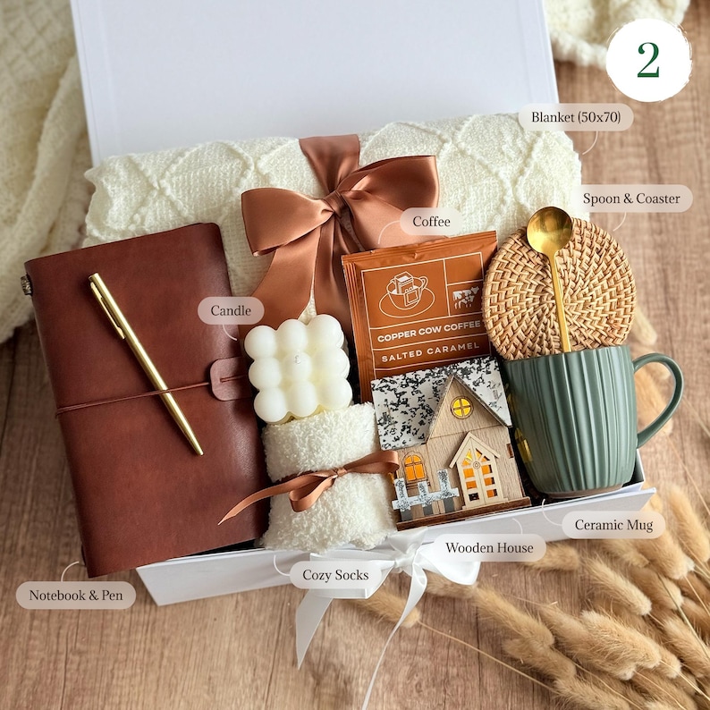 May include: A gift box filled with a cream-colored blanket, a brown leather notebook and pen, a white candle, a packet of coffee, a wooden house, a green ceramic mug, a woven coaster with a gold spoon, and cozy socks. The box is tied with a brown ribbon.