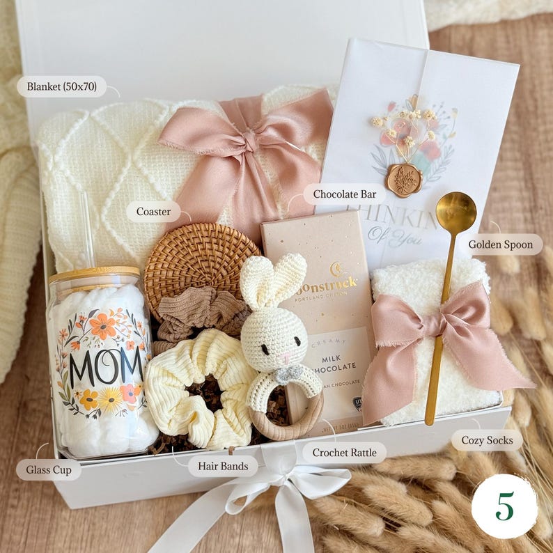 May include: A gift box filled with a cream-colored blanket (50x70 inches), a glass cup with "MOM" text, a crochet rattle, a chocolate bar, a golden spoon, and other items. The box is tied with a white ribbon.