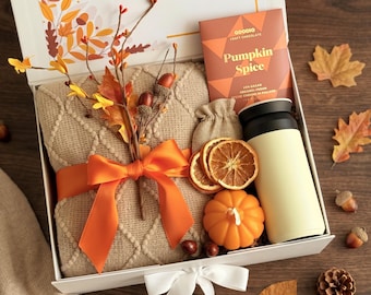 May include: A gift box containing a beige knit blanket tied with an orange ribbon, a Goodio Pumpkin Spice chocolate bar, a cream-colored travel mug, a small orange pumpkin candle, and dried orange slices, with text reading AND MARCUS.
