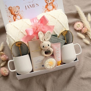 Pu&ograve; includere: Un cesto regalo con articoli a tema baby. Include una coperta bianca, due tazze bianche, un sonaglio a forma di coniglietto all'uncinetto, una scatola di cioccolato al latte, un tubetto di lozione alla rosa e un fiore decorativo. La scatola &egrave; decorata con orsetti e i nomi "and James".