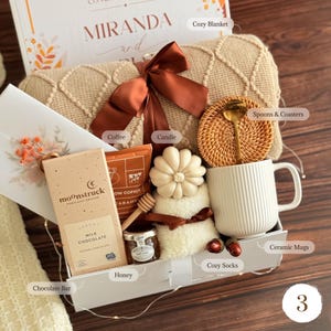 May include: A gift box containing a beige knit blanket with a brown ribbon, a Moonstruck milk chocolate bar, a bag of coffee, a flower-shaped candle, a jar of honey, a wooden honey dipper, a white ribbed ceramic mug, and a rattan coaster with a gold spoon.