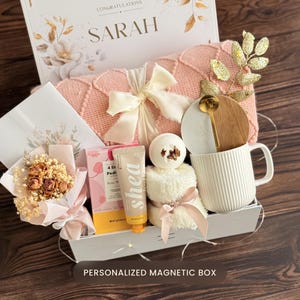 May include: A gift box with a pink blanket, a white mug, a bouquet of dried flowers, and a tube of shea butter lotion. The box also contains a bath bomb, a wooden coaster with a gold spoon, and a card that says "Congratulations Sarah". The box is labeled "Personalized Magnetic Box".