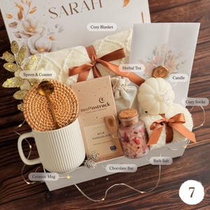 May include: A gift box containing a white ribbed ceramic mug with a rattan coaster and gold spoon, a Moonstruck milk chocolate bar, a glass jar of bath salts, a white pumpkin-shaped candle, a cream knit blanket, and herbal tea with a wax seal.