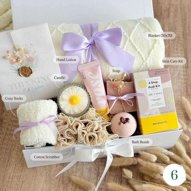 May include: A gift box filled with self-care items. Includes a cream-colored blanket (50x70 inches), cozy socks, a candle, hand lotion, soap, a bath bomb, a skin care kit, and a cotton scrubber. The box is tied with a white bow and a lavender ribbon.