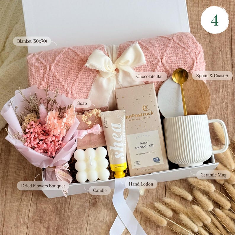 May include: A gift box filled with self-care items, including a pink blanket, a white ceramic mug, a bar of chocolate, a hand lotion, a candle, a bar of soap, a dried flower bouquet, and a spoon and coaster.