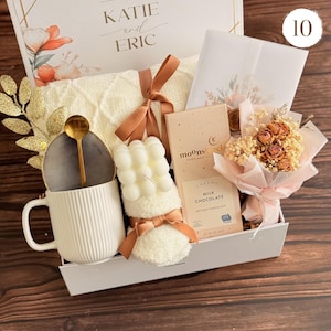 Peut inclure: Un coffret cadeau avec une tasse blanche, une cuill&egrave;re dor&eacute;e, une bougie, une barre de chocolat et un petit bouquet. La bo&icirc;te comprend une couverture cr&egrave;me, une carte et une composition florale d&eacute;corative. La bo&icirc;te est &eacute;tiquet&eacute;e "KATIE and ERIC".
