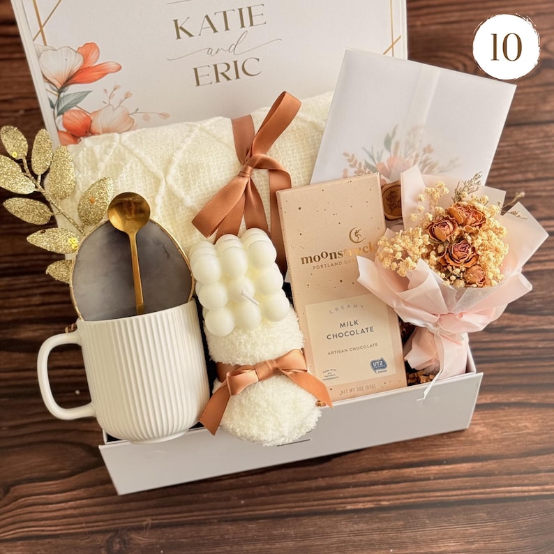Peut inclure: Un coffret cadeau avec une tasse blanche, une cuill&egrave;re dor&eacute;e, une bougie, une barre de chocolat et un petit bouquet. La bo&icirc;te comprend une couverture cr&egrave;me, une carte et une composition florale d&eacute;corative. La bo&icirc;te est &eacute;tiquet&eacute;e "KATIE and ERIC".