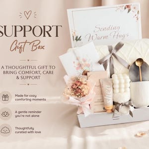 May include: A gift box containing a white ribbed mug with a gold spoon, a bubble candle, a tube of shea cream, a milk chocolate bar, a small dried flower bouquet, a white knit blanket with a grey ribbon, and a card reading Sending Warm Hugs.