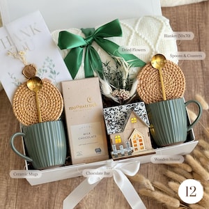 May include: A gift box with a white blanket, green ceramic mugs, a chocolate bar, and a wooden house. The box includes a green bow, dried flowers, spoons, and coasters. The blanket measures 50x70 inches.