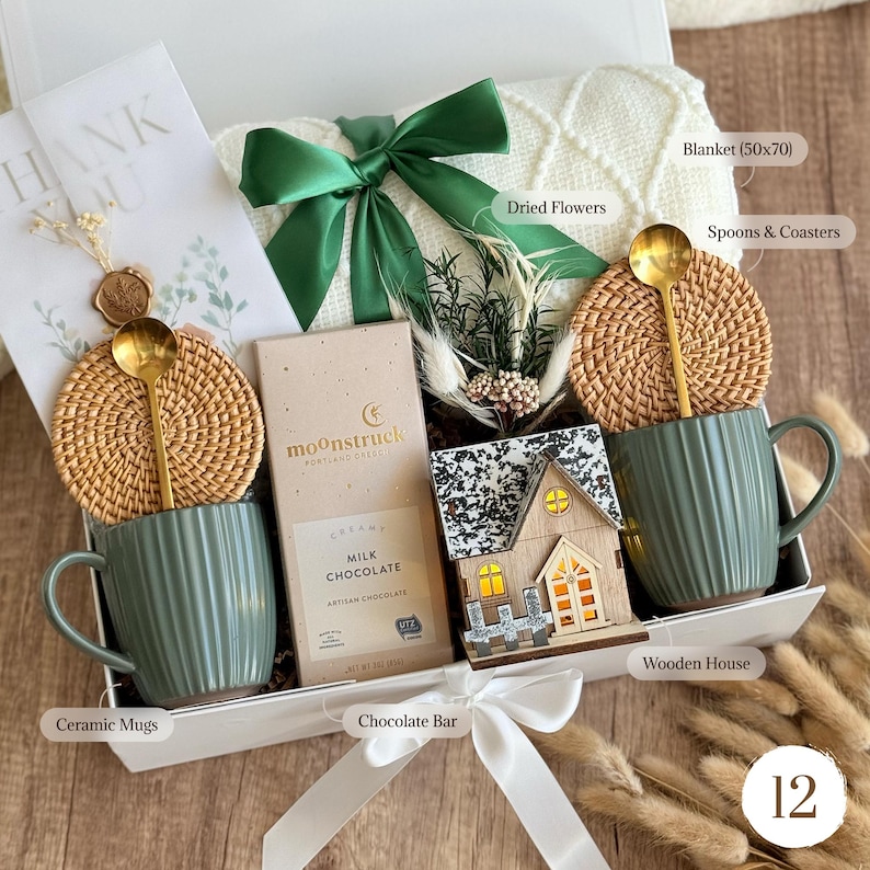 May include: A gift box with a white blanket, green ceramic mugs, a chocolate bar, and a wooden house. The box includes a green bow, dried flowers, spoons, and coasters. The blanket measures 50x70 inches.