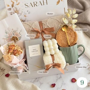 May include: A gift box with a beige blanket, a ceramic mug, a candle, hot cocoa, cozy socks, a dried flower bouquet, and a spoon on a coaster. The box is decorated with floral designs and the name "SARAH".
