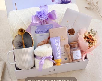 May include: A gift box containing a white ribbed mug with a gold spoon, a pair of lavender shea gloves, a white textured blanket, a small towel tied with a purple ribbon, a lavender hand cream, a chocolate bar, bath salts, and a small dried flower bouquet.