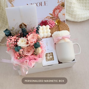 May include: A gift box containing a bouquet of pink and white preserved roses, a white bubble candle, a white ribbed ceramic mug with a pink ribbon, a gold spoon, a rattan coaster, and a box of champagne bubbles candy with a floral lid.