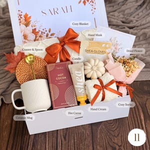 May include: A gift box containing a white ceramic mug, a gold spoon, a woven coaster, a box of hot cocoa, a shea butter hand cream, a shea butter hand mask, a pumpkin-shaped candle, white fuzzy socks with a ribbon, and a small dried flower bouquet.