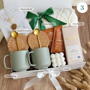 May include: A white gift box with a green ribbon and a white blanket. The box contains two ceramic mugs, two woven coasters, two gold spoons, two packets of coffee, a chocolate bar, a candle, and a small white soap. The box is labeled with the text "Coffee for Two", "Chocolate Bar", "Spoons & Coasters", "Ceramic Mugs", "Blanket (50x70)", and "Candle".