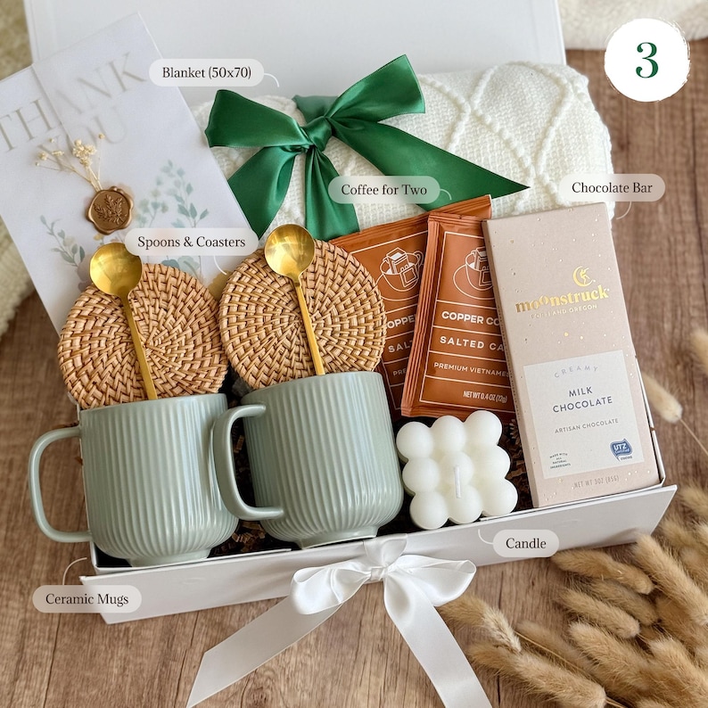 May include: A white gift box with a green ribbon and a white blanket. The box contains two ceramic mugs, two woven coasters, two gold spoons, two packets of coffee, a chocolate bar, a candle, and a small white soap. The box is labeled with the text "Coffee for Two", "Chocolate Bar", "Spoons & Coasters", "Ceramic Mugs", "Blanket (50x70)", and "Candle".