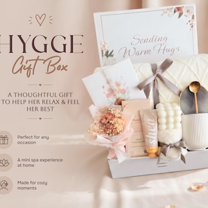 May include: A Hygge gift box with a white blanket, mug, and other spa items. The box includes a bouquet of flowers, a tube of Shea lotion, and a card that says "Sending Warm Hugs."