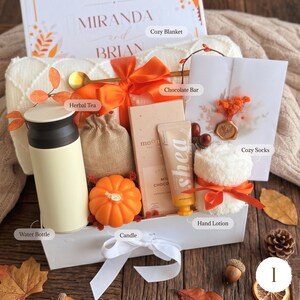 May include: A gift box containing a cream water bottle, a burlap pouch of herbal tea, an orange pumpkin-shaped candle, a chocolate bar, a tube of shea hand lotion, and white cozy socks tied with an orange ribbon, all set against a white blanket.