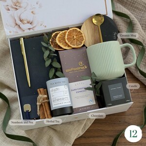 May include: A gift box containing a black notebook with a gold pen, a light green ribbed ceramic mug, a gold spoon, a dark chocolate bar, a tin of herbal tea, a candle, dried orange slices, and cinnamon sticks, all arranged in a white box.