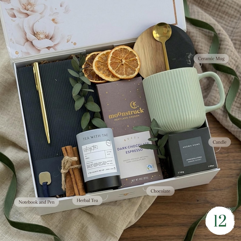 May include: A gift box containing a black notebook with a gold pen, a light green ribbed ceramic mug, a gold spoon, a dark chocolate bar, a tin of herbal tea, a candle, dried orange slices, and cinnamon sticks, all arranged in a white box.