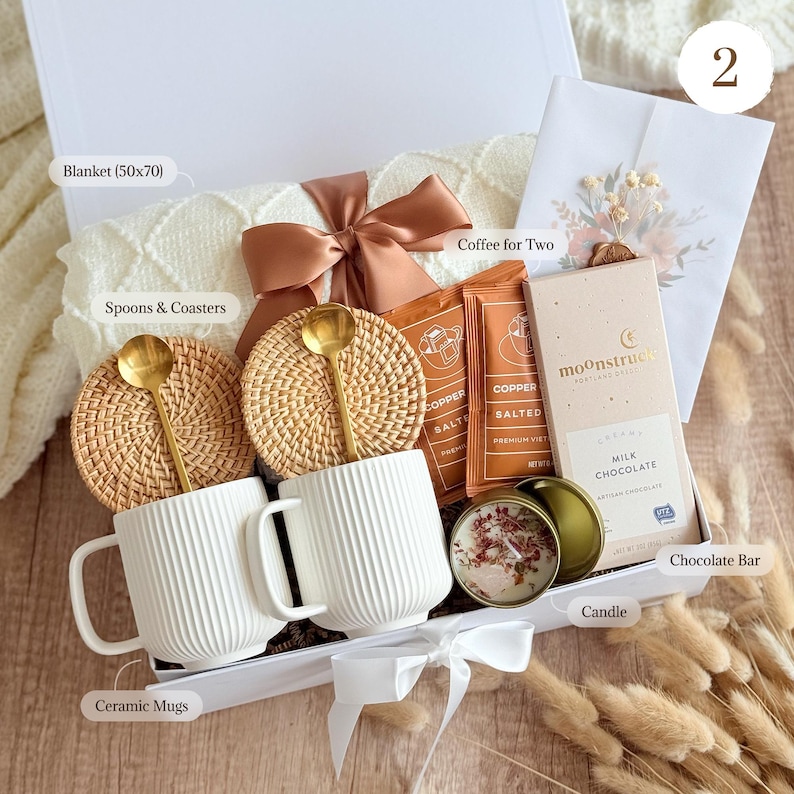 May include: A gift box containing two white ribbed ceramic mugs, two gold spoons, two round woven coasters, a white knit blanket with a copper ribbon, two coffee packets, a Moonstruck milk chocolate bar, and a floral candle in a gold tin.
