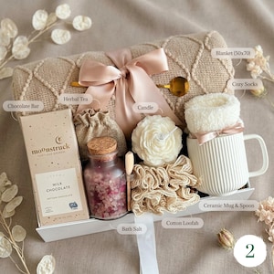 May include: A gift basket with a beige blanket (50x70 inches) tied with a pink ribbon. It includes a chocolate bar, herbal tea, a candle, bath salts, a ceramic mug with cozy socks, and a cotton loofah.
