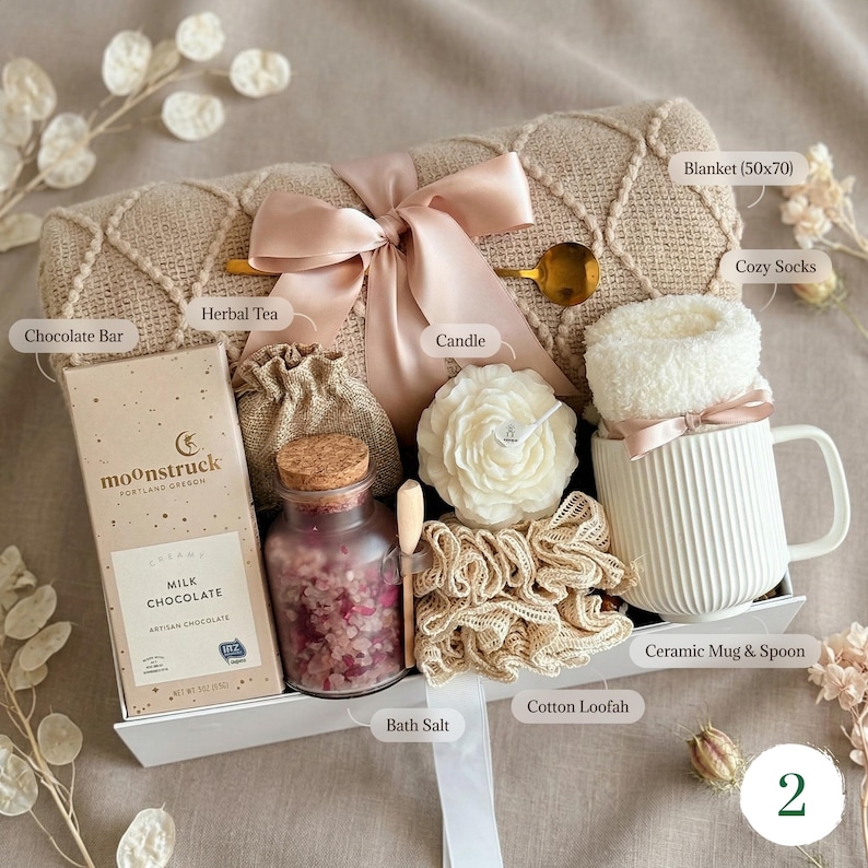 May include: A gift basket with a beige blanket (50x70 inches) tied with a pink ribbon. It includes a chocolate bar, herbal tea, a candle, bath salts, a ceramic mug with cozy socks, and a cotton loofah.