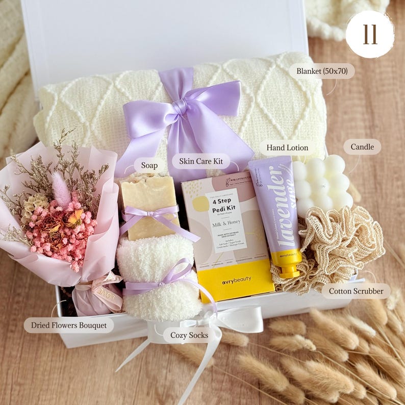 May include: A white gift box filled with self-care items. Includes a cream-colored blanket (50x70 inches), lavender-scented hand lotion, a candle, a skin care kit, soap, a dried flower bouquet, cozy socks, and a cotton scrubber.