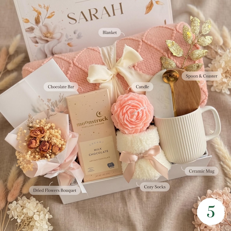 May include: A gift box containing a pink blanket, a white ceramic mug, a rose-shaped candle, a chocolate bar, a bouquet of dried flowers, cosy socks, and a wooden spoon. The box is labelled "SARAH".
