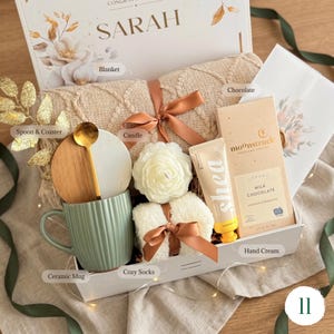 May include: A gift box with a blanket, candle, mug, socks, chocolate, hand cream, spoon, and coaster. The box is decorated with a ribbon and the name "SARAH". The items are in neutral colors with a floral design.