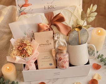 May include: A gift box with a cream-colored blanket, a mug, chocolate, a small bouquet, and a candle. The box is decorated with a bow and the words "Happy Engagement" and the names "Katie and Eric". The mug has a gold spoon and a decorative stone.