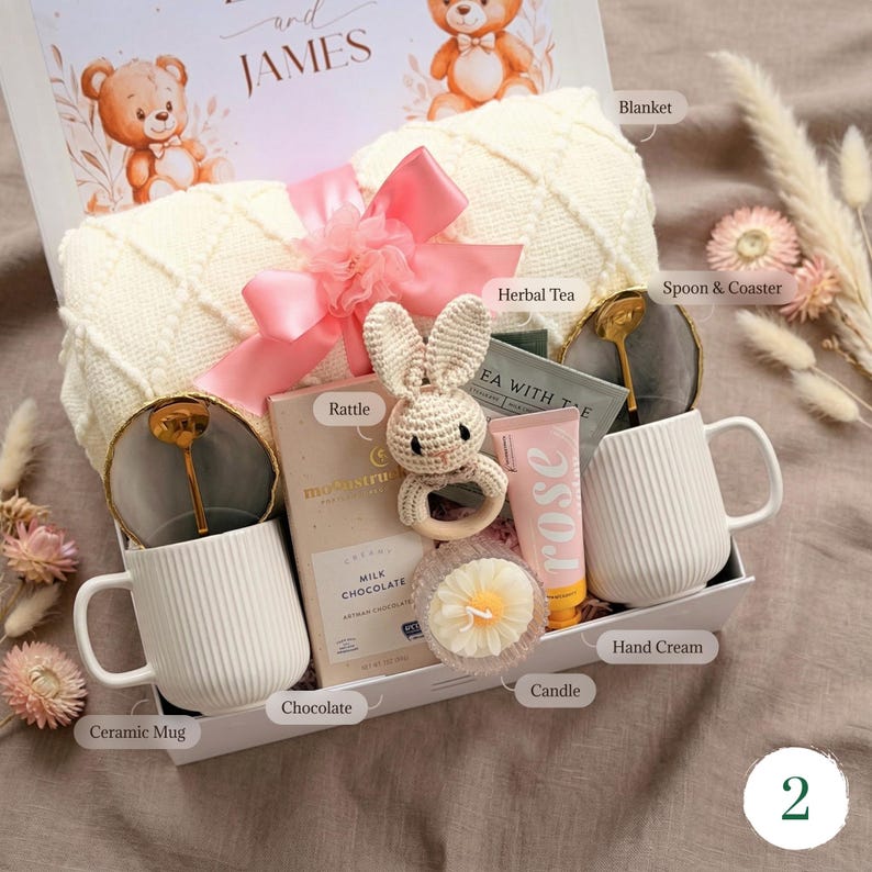 Pu&ograve; includere: Una confezione regalo con una coperta color crema, un fiocco rosa e vari articoli. Include una tazza in ceramica, un sonaglio, cioccolato, crema per le mani, una candela e tisana. La scatola &egrave; decorata con orsetti e il nome "JAMES".