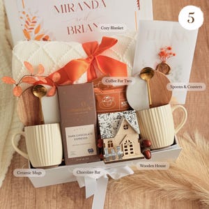 May include: A gift box with a cozy blanket, ceramic mugs, chocolate bar, coffee, wooden house, spoons, and coasters. The box is tied with an orange ribbon. The box includes a card that says "MIRANDA and BRIAN".