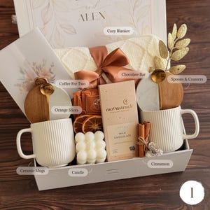 May include: A gift box containing two white ribbed ceramic mugs, a Moonstruck milk chocolate bar, a white bubble candle, dried orange slices, cinnamon sticks, two gold spoons, two round coasters, and a cream-coloured knit blanket with a brown ribbon.