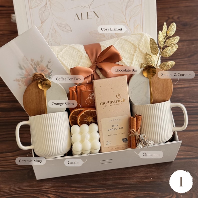 May include: A gift box containing two white ribbed ceramic mugs, a Moonstruck milk chocolate bar, a white bubble candle, dried orange slices, cinnamon sticks, two gold spoons, two round coasters, and a cream-coloured knit blanket with a brown ribbon.