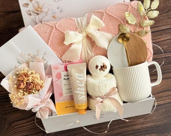 Puede incluir: Una caja de regalo con una manta de punto color melocotón con lazo crema, una taza blanca acanalada con tapa de madera y cuchara dorada, un ramo de flores secas, un tubo de manteca de karité, una bomba de baño y una toalla blanca con lazo.